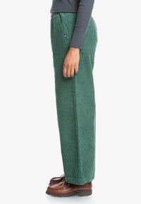 Corduroy pants in dark green with a straight leg design, featuring front pockets and a visible brand tag on the waistband. Brown shoes.