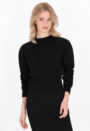 Pullover nero con un collo alto, tessuto strutturato e decorazioni di perline sulle spalle, abbinato a una gonna nera aderente.