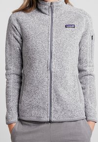 Grå fleecjacka med hög krage och dragkedja framtill. Har sidofickor och en liten logo på bröstet. Slät textur, figurnära design.