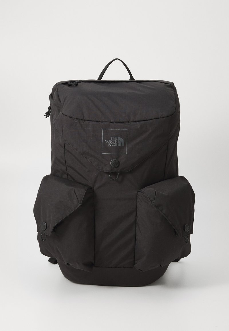 Mochila de nylon preta com um topo arredondado, alças de ombro ajustáveis, bolsos frontais e um patch de logotipo. Possui fechos de correr e botões de pressão.