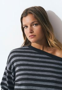 Suéter a rayas en gris y negro, hecho de un suave tejido de punto con un ajuste holgado. Presenta un escote asimétrico y sutiles toques de brillo.