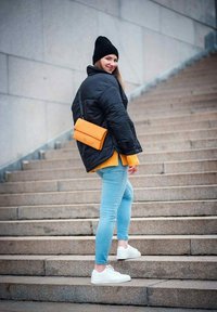 Černá péřová bunda přes jasně žlutý svetr, světle modré džíny, bílé tenisky a oranžová kabelka. Model pózuje na kamenných schodech.