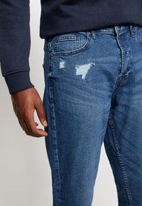 Calças de ganga azuis com um corte slim, ligeiramente desgastadas na coxa, e um clássico design de cinco bolsos, combinadas com uma camisa de manga longa escura.