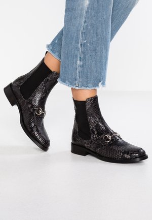 Person iført sorte skinnende ankelstøvler med slangeskindsmønster, sølvspænde og blå jeans med frynset kant.