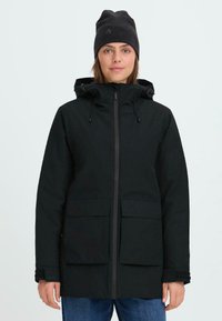 Chaqueta aislante negra con capucha, cierre de cremallera, grandes bolsillos frontales y puños ajustables. Llevada con un gorro negro a juego.