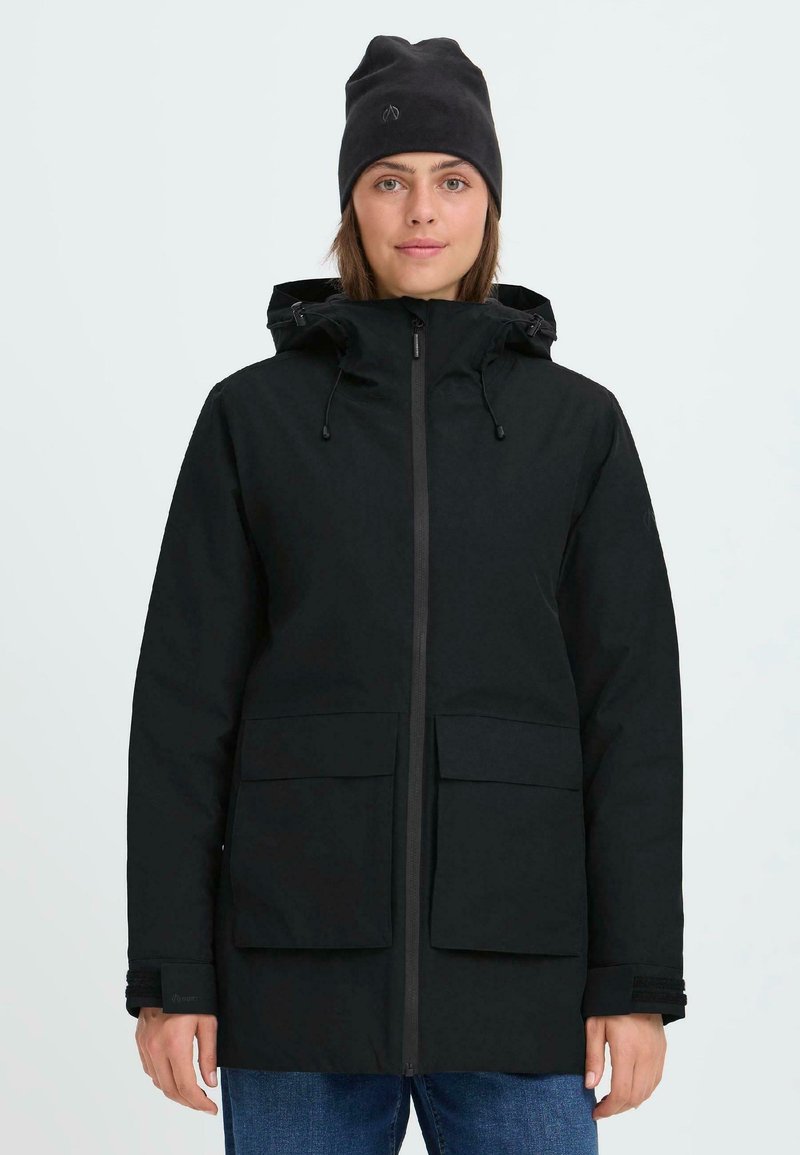 Chaqueta aislante negra con capucha, cierre de cremallera, grandes bolsillos frontales y puños ajustables. Llevada con un gorro negro a juego.