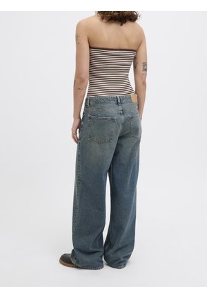 Persona con cabello rizado, vistiendo un top tipo tubo a rayas marrones y blancas y pantalones vaqueros azules holgados, de pie de espaldas sobre un fondo liso.