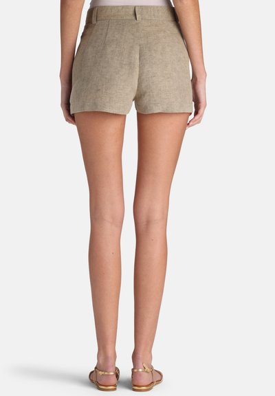 Femme portant un short en lin beige et des sandales plates, debout de dos sur un fond uni.