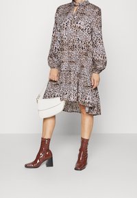 Šaty s leopardím potiskem a dlouhými nafouknutými rukávy, volný střih a stupňovitý lem; zkombinováno s lesklými hnědými kotníkovými botami na podpatku a bílou kabelkou.