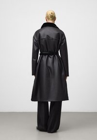 Zwarte lange jas met een ceintuur in de taille, gemaakt van glanzend materiaal, met een zachte bontkraag en een eenvoudig ontwerp. Draagt over wijde broekspijpen.