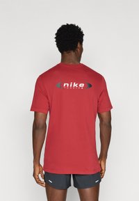 Κόκκινο μπλουζάκι Nike με κοντά μανίκια και στρογγυλή λαιμόκοψη, με μαύρο λογότυπο στο πίσω μέρος. Το ύφασμα φαίνεται λείο και ελαφρύ.