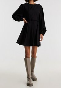 Robe noire à manches longues avec une légère silhouette trapèze, ceinture à la taille et détail d'encolure en forme de goutte, associée à des bottes en caoutchouc grises montantes jusqu'aux genoux.