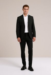 Fekete öltöny, testhez simuló dizájnnal, egysoros gombos zakóval és klasszikus hajtókával. Fehér inggel és fekete fűzős cipővel párosítva.