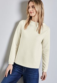 Helles gelbes gestepptes Sweatshirt mit Rundhalsausschnitt und langen Ärmeln, kombiniert mit dunkelblauen Jeans. Die Textur weist ein Rautenmuster auf.