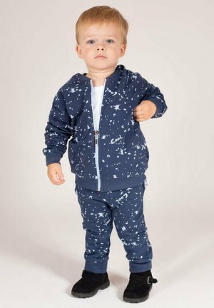 Granatowy dziecięcy dres z białym wzorem rozprysków, kurtka na zamek, ściągacze przy rękawach oraz pasujące spodnie, w komplecie z czarnymi butami.