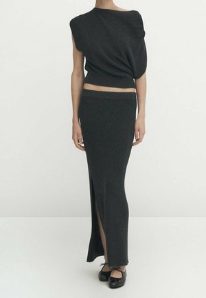 Femme portant un haut asymétrique sombre sans manches et une longue jupe assortie avec une fente à l'avant, associée à des ballerines noires.