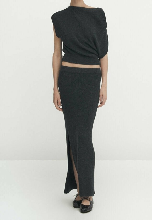 Femme portant un haut asymétrique sombre sans manches et une longue jupe assortie avec une fente à l'avant, associée à des ballerines noires.