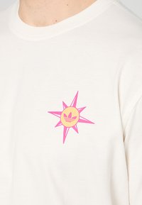 T-shirt color crema con collo rotondo, caratterizzato da un vivace design di una stella rosa e un cerchio giallo con un logo al centro.
