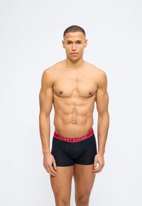 Μυώδης άνδρας όρθιος, στραμμένος προς τα εμπρός, φοράει μαύρα μπόξερ Tommy Hilfiger με κόκκινη μέση, σε απλό λευκό φόντο.