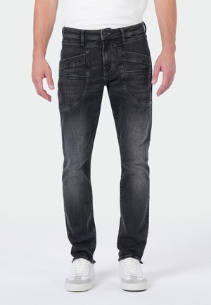 Man wearing faded black jeans with multiple front pockets and white sneakers, standing against a plain background.