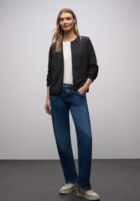 Czarna pikowana kurtka bomberka z okrągłym dekoltem, zestawiona z niebieskimi szerokimi spodniami i kremowymi sneakersami, stojąca na neutralnym tle.