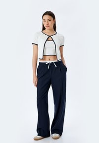 Witte cropped top met marineblauwe biezen, met een open voorkant. Marineblauwe wijde broek met een trekkoord in de taille. Licht materiaal en relaxed pasvorm.