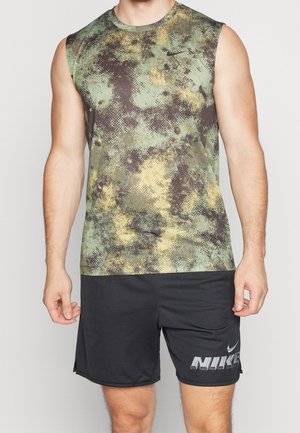 Man met een groene en gele camouflagenike-singlet zonder mouwen en zwarte Nike-short met logo op linkerbeen, staand tegen een effen achtergrond.
