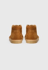 Stivali alla caviglia in suede marrone con punta arrotondata, colletto corto, cuciture visibili e suola in gomma leggera. La texture è morbida e opaca.