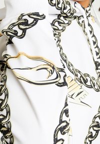 Bílé mikina s kapucí s černým a zlatým potiskem řetězu a částečně viditelným logem Nike na hrudi.