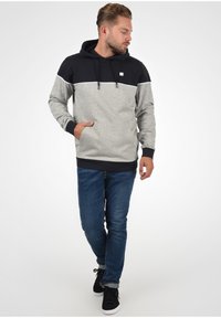 Hoodie met een zwart bovenstuk en een grijs lichaam, voorzien van een witte streep. Kangaroo zak, geribde manchetten en zoom. Gecombineerd met een blauwe spijkerbroek.