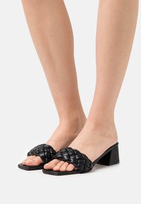 Des pieds portant des sandales à talons en cuir tressé noir avec des orteils carrés sur fond blanc.