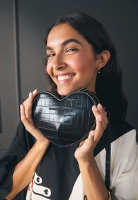 Melnas sirds formas soma no teksturētas mākslīgās ādas ar krokodila rakstu, rāvējslēdzēja aizdari un plecu siksnu. Zīmola nosaukums ir reljefā.