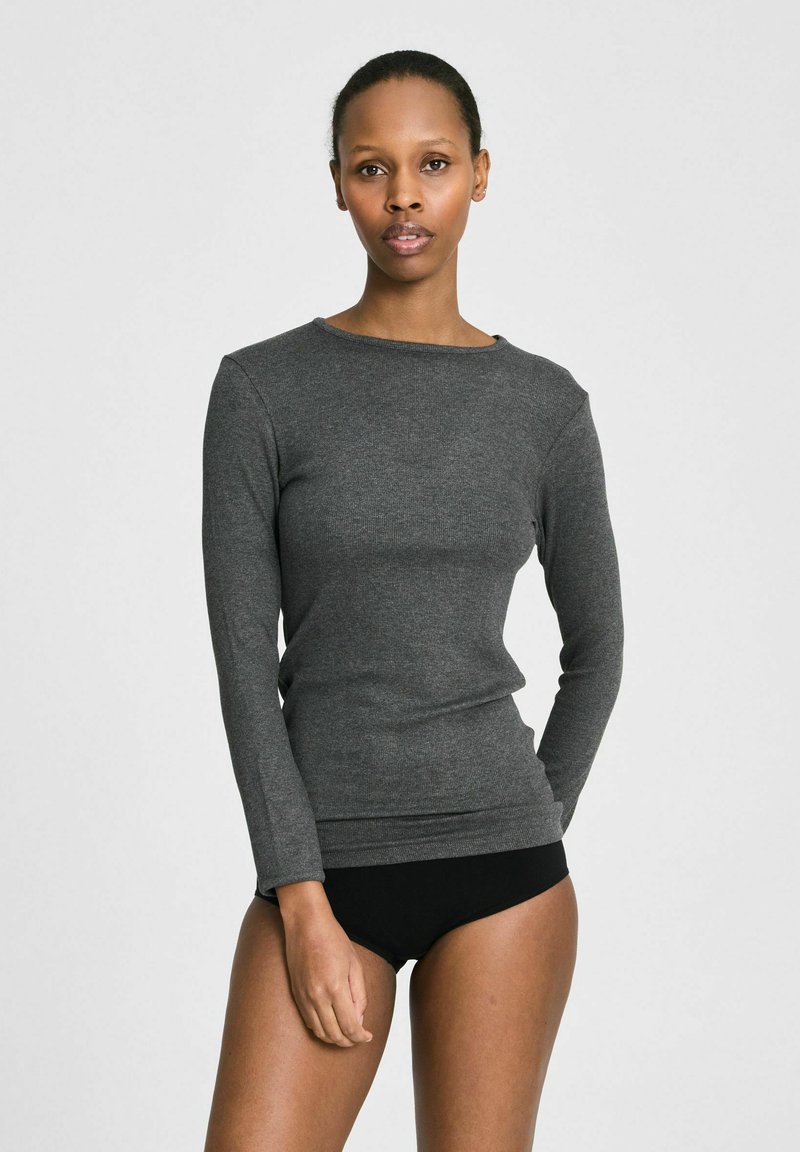 Femme portant un haut gris ajusté à manches longues et des sous-vêtements noirs, debout devant un fond uni gris clair, faisant face à l'avant.