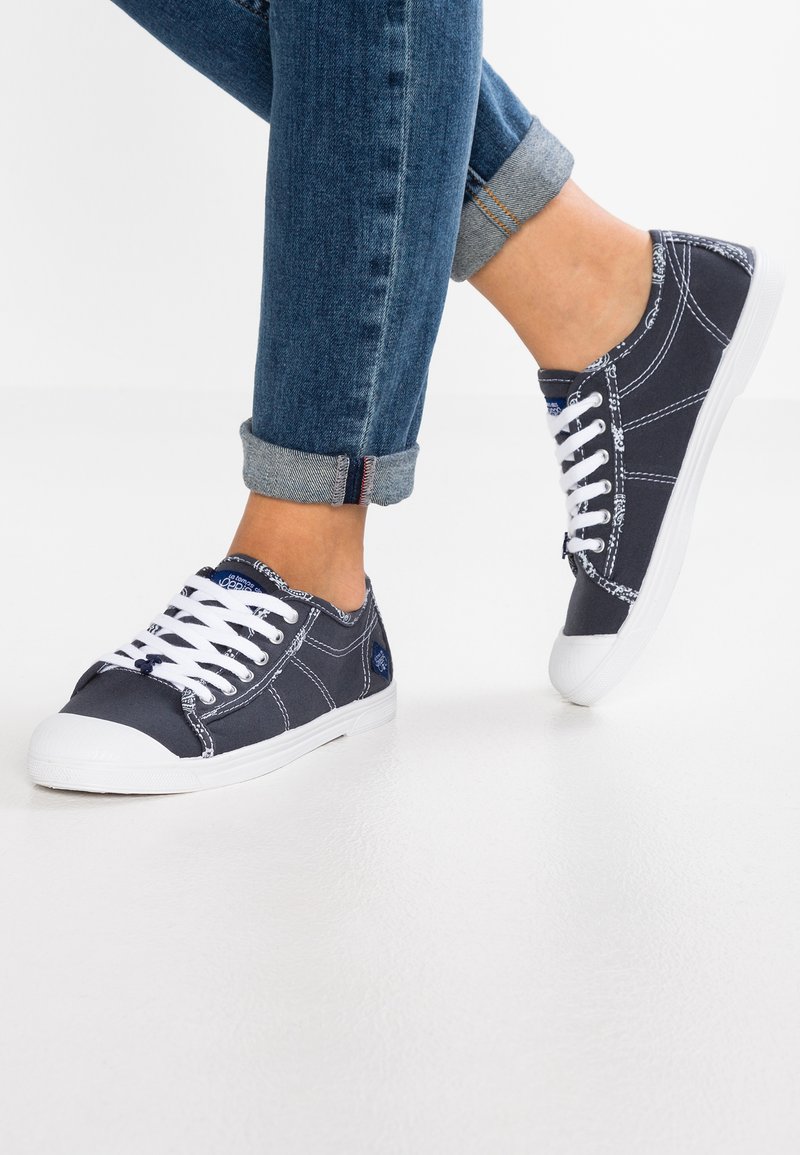 Sneakers di tela blu scuro con lacci bianchi, suole in gomma e cuciture in contrasto. Degli jeans in denim risvoltati sono parzialmente visibili.
