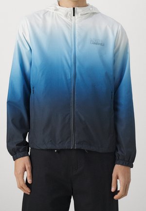 Homme portant une veste à capuche zippée avec un dégradé bleu-noir, des poignets élastiques, des poches latérales, et le logo "KARL LAGERFELD" sur la poitrine.
