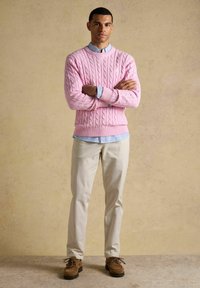 Man staat met gekruiste armen, draagt een roze kabelgebreide trui, een lichtblauw gestreept overhemd, beige broek en bruine schoenen tegen een neutrale muur.