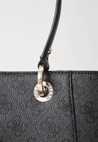 Musta tekstuuriga käekott, millel on õhuke must käepide, mille küljes on kuldne rõngas, millele on graveeritud "GUESS", sileda tausta peal.