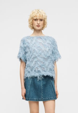 Jong persoon met kort krullend blond haar, gekleed in een doorschijnende lichtblauwe top met textuur en een spijkerbroekminirok, tegen een effen achtergrond.