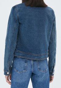 Veste en denim avec un lavage foncé, présentant un design simple, des manches longues, des poignets à boutons et des coutures visibles. Associée à un jean en denim assorti.