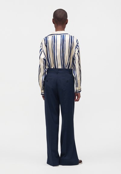 Personne vue de dos portant une chemise crème à rayures verticales bleu marine et un pantalon large taille haute bleu marine, debout devant un fond uni.