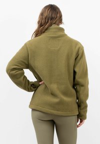 Olivgrüne Fleecejacke mit hohem Kragen, strukturiertem Stoff und eingesetzten Ärmeln. Trägt dazu passende, enganliegende Hose. Keine sichtbaren Verschlüsse.