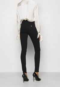 Jeans negros ajustados de denim, combinados con una blusa de satén beige y zapatos de tacón negros. Detalle de logo visible en el bolsillo trasero.