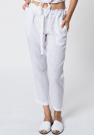 Kobieta w białych lnianych spodniach ze ściągaczem i białych wsuwanych butach, stojąca z rękami w kieszeniach na jednolitym tle.