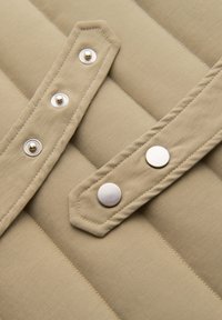 Tela acolchada beige con dos botones a presión en una correa y tres tachuelas metálicas en una correa paralela, mostrando una costura detallada.