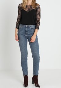 Top nero in pizzo a maniche lunghe con torso trasparente su una base nera solida, abbinato a jeans blu a vita alta e stivaletti a punta color bordeaux.