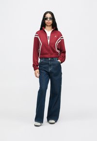 Czerwona kurtka zapinana na zamek z białymi paskami, zestawiona z szerokimi niebieskimi dżinsami i szarymi trampkami. Model ma na sobie okulary przeciwsłoneczne i białą koszulkę z grafiką.