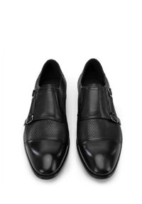 Zapatos de piel negra con una punta lisa y un patrón herringbone texturizado en la parte superior, con dos hebillas de metal a los lados.