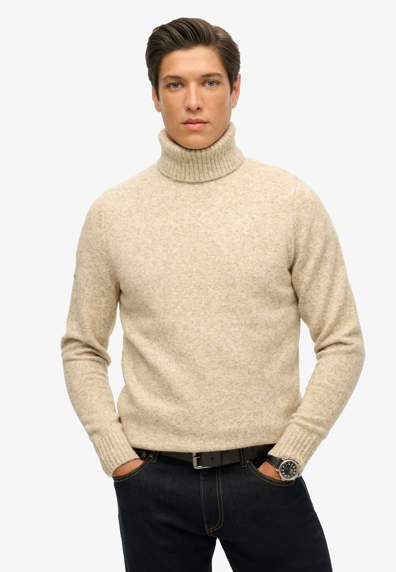 Suéter de cuello alto beige de punto con puños y dobladillo acanalados, combinado con jeans oscuros y un cinturón negro. La textura parece suave y cálida.