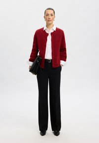 Cardigan de lana roja con cierre de botón sobre una camisa blanca con cuello, combinado con pantalones de pierna ancha negros y zapatos de tacón negros. Incluye un bolso de mano negro.