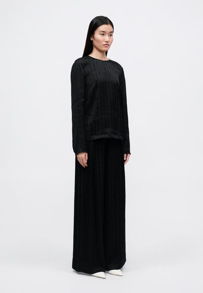 Svart langermet topp og brede bukser i strukturert stoff; begge har et subtilt mønster. Antrekket opprettholder en minimalistisk design.